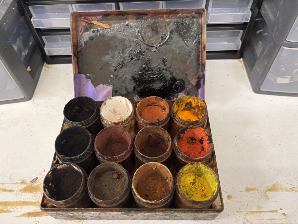 Vue de dessus d'une boîte en métal ancienne ouverte, contenant douze petits pots en verre remplis de pigments en poudre de différentes couleurs (terres, ocres, rouges, jaunes, noirs) pour la restauration de mobilier.