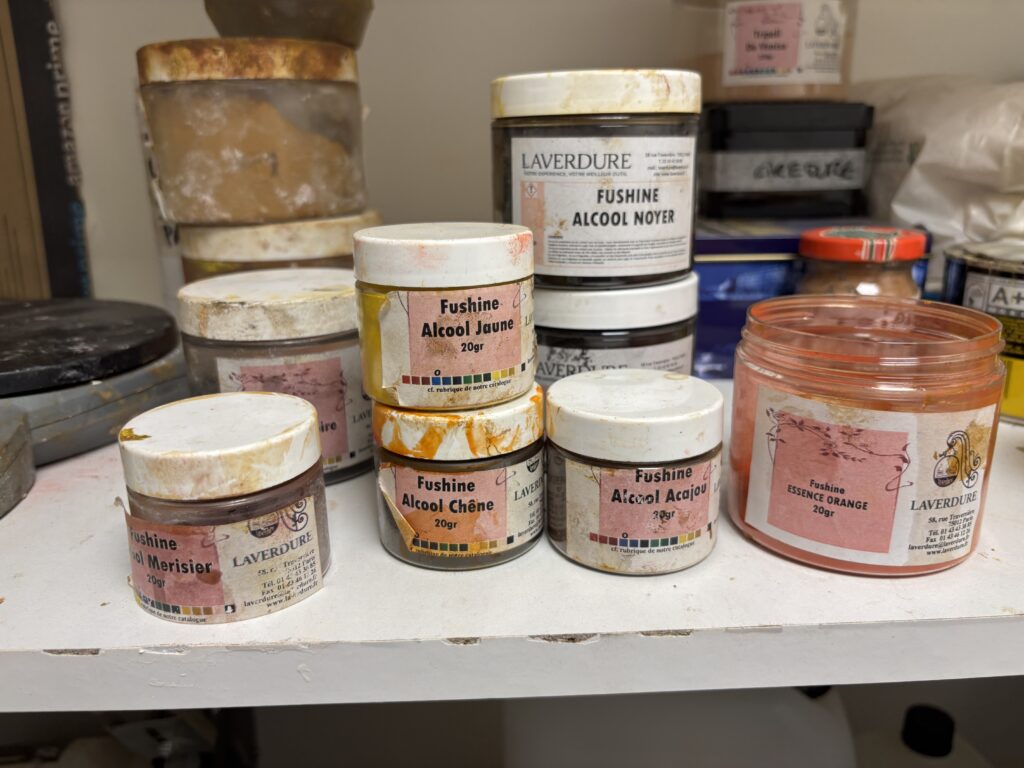 Gros plan sur plusieurs petits pots de marque Laverdure alignés sur une étagère d'atelier. Les étiquettes indiquent "Fushine Alcool" avec des nuances comme Noyer, Jaune, Acajou, Chêne, Merisier, Orange.