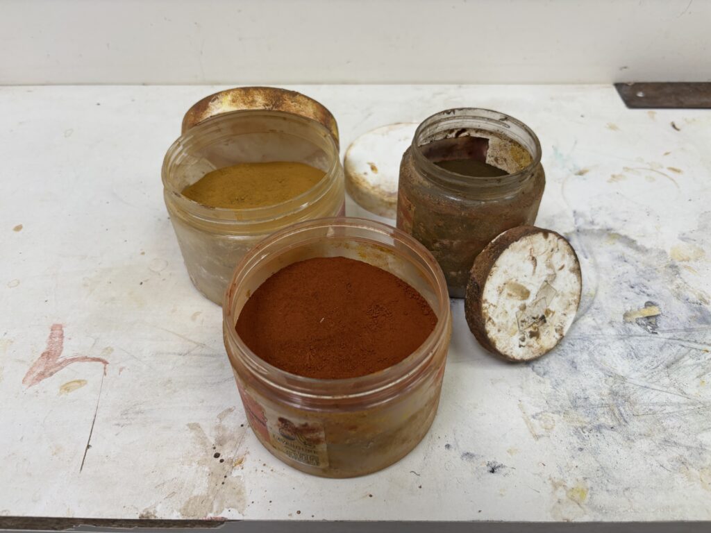 Trois pots en plastique transparent ouverts sur un plan de travail d'ébéniste taché. Ils contiennent des pigments en poudre en gros plan : un ocre jaune vif, un rouge sienne profond et une terre d'ombre.